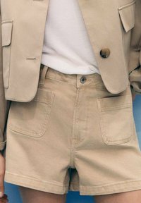 Pantalones cortos de mezclilla beige con dos bolsillos delanteros y cierre de botón. Los pantalones cortos tienen un corte recto y se combinan con una blusa clara y un blazer.