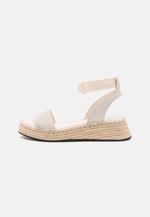 Beige platformsandal med vævet jutesål, åben tårem og justerbar ankelrem med velcrolukning på hvid baggrund.