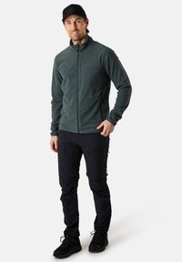 Fleece-zipattava takki tummanvihreänä, istuva malli taskuilla; yhdistetty mustiin cargo-housuihin, joissa on vetoketjuja ja liukutaskuja, viimeistelty mustilla lenkkareilla.