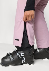 Pantaloni de schi roz cu căptușeală neagră și deschideri cu fermoar la picioare, purtați peste cizme de schi negre cu marcaj alb și catarame din metal.