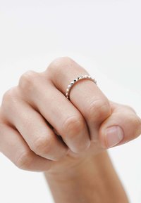 Anillo de plata con una banda redondeada que presenta pequeños picos triangulares a lo largo del borde superior. Llevarlo en una mano de tono de piel claro.