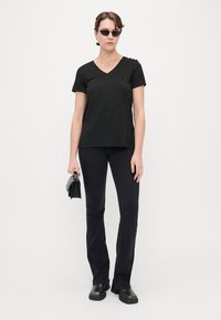 Sort v-hals t-shirt med en tekstureret finish, parret med sorte flared bukser og kraftige sorte sko. Modellen holder en lille sort taske.