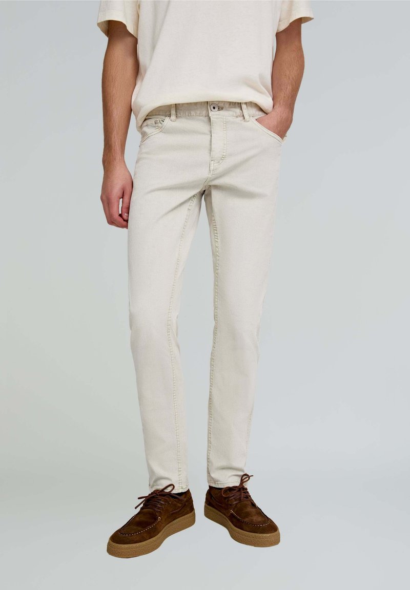 Beige Baumwolljeans mit schlankem Schnitt, klassischer Fünf-Taschen-Style und dezenten Nähdetails. Kombiniert mit braunen Wildlederschuhen.