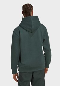 Person med flätat hår som bär en mörkgrön huvtröja och matchande byxor, sedd bakifrån mot en enfärgad bakgrund.