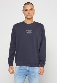 Donkerblauwe lange mouw sweatshirt van katoen, met een ronde halslijn en witte bedrukte tekst op de borst. Draagt met lichtblauwe spijkerbroek.
