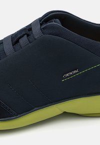 Scarpa blu navy con dettagli in rete, caratterizzata da una parte superiore liscia e una suola verde a contrasto. Logo presente sul lato con sottili accenti di cucitura.