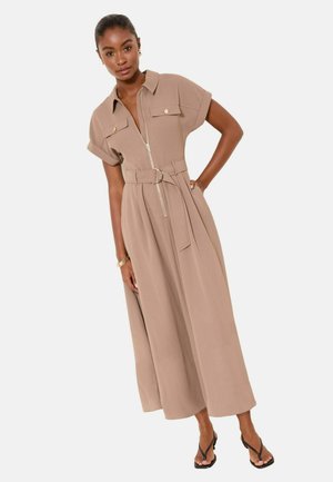 Frau trägt ein beige-gürtelt Midikleid mit kurzen Ärmeln und einem Frontreißverschluss, kombiniert mit schwarzen Riemchensandalen und einem goldenen Armband.