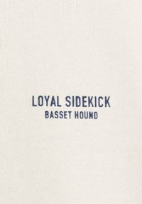 Tkanina boje kreme s rebrastom teksturom koja sadrži tamnoplavi tekst: "LOYAL SIDEKICK" i "BASSET HOUND" u podebljanom, jednostavnom fontu.