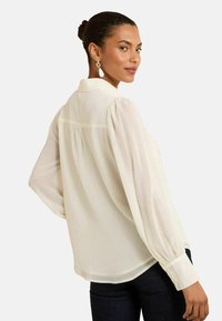 Blusa cremosa a manica lunga con colletto, tessuto strutturato, yoke posteriore arricciato e polsini con bottoni. Indossata con jeans scuri.