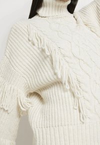 Pull en tricot blanc avec un col haut, présentant une texture côtelée et des bords à franges, ainsi qu'un motif torsadé sur le devant.