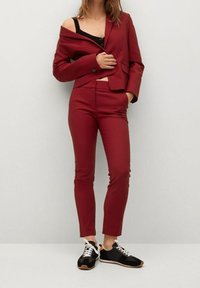 Blazer sur mesure de couleur cramoisie avec une épaule découverte, pantalon slim assorti, et baskets noires avec lacets blancs et semelles en gomme.