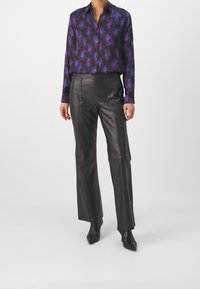 Lila, floral gemusterte Bluse mit langen Ärmeln, kombiniert mit hochgeschnittenen schwarzen Lederhosen. Das Outfit zeichnet sich durch ein elegantes, figurnahes Design und spitze Stiefel aus.