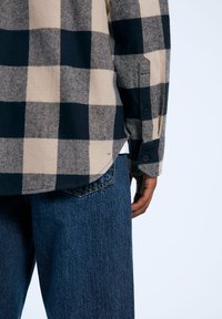 Camisa de cuello con un patrón de cuadros en beige y azul marino, fabricada con una tela suave, con puños abotonados y un dobladillo curvado. Llevada con pantalones de mezclilla azul.