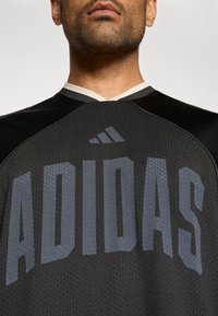 Czarny sportowy dres wykonany z perforowanej tkaniny, z szarym logo "ADIDAS" na przodzie oraz kontrastującym jasnym kołnierzykiem.