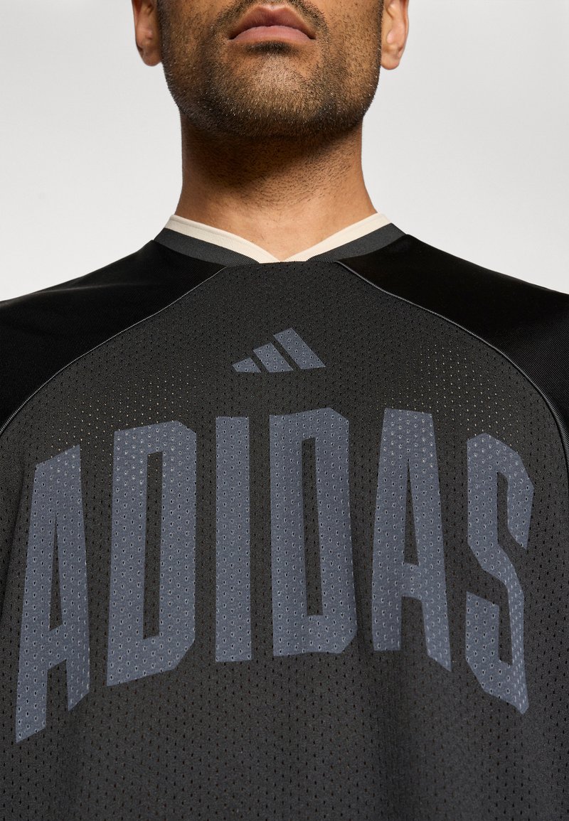 Svart sporttröja av perforerat tyg med en grå "ADIDAS"-logotyp över framsidan och en kontrasterande ljusfärgad halsringning.