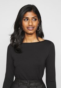 Black long-sleeve top with a boat neckline, paired with high-waisted black leather pants. The model has straight black hair and hoop earrings.