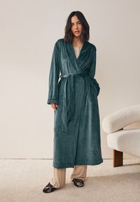 Dlhý, tyrkysový plyšový župan s prekladaným strihom, opaskom a bočnými vreckami, doplnený svetlo béžovými nohavicami a vzorovanými papučami. Mäkká textúra.