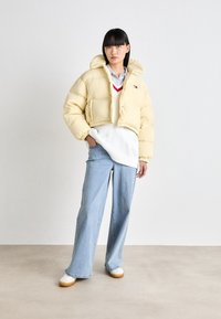 Geel cropped pufferjack met een capuchon, voorzien van een rits aan de voorkant en elastische manchetten. Draagt boven een witte trui en blauwe wijde jeans.
