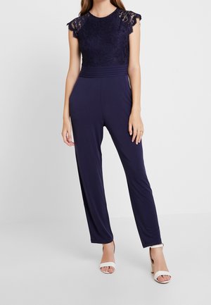 Kvinde iført en navy buksedragt med blonder på kappelærmer og plisseret taljebånd, kombineret med hvide åbne hælede sandaler.