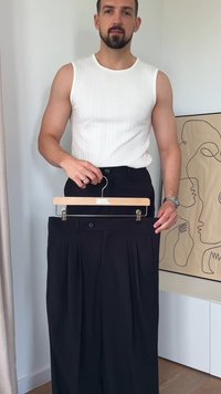 Black pleated trousers with front pockets, hung on a wooden hanger. High-waisted design, minimalistic style. Smooth fabric texture.