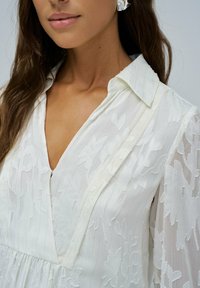 Blusa bianca con ricami floreali, scollo a V e colletto. Presenta un tessuto leggero e trasparente con strisce testurizzate e una vestibilità comoda.