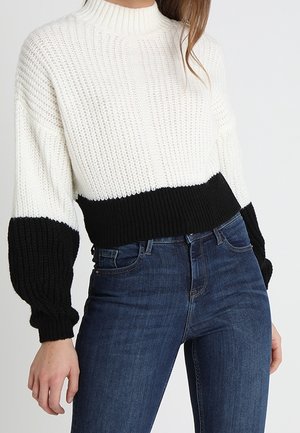 Femme portant un pull en maille blanc avec des poignets et un ourlet noirs, associé à un jean taille haute bleu foncé sur un fond uni.