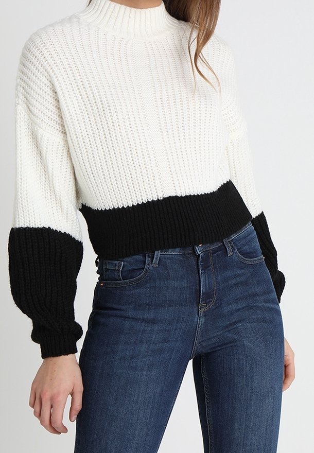 Femme portant un pull en maille blanc avec des poignets et un ourlet noirs, associé à un jean taille haute bleu foncé sur un fond uni.