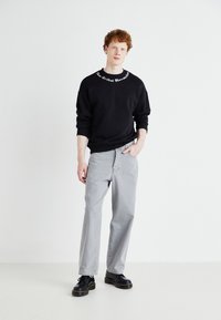 Black sweatshirt with white embroidered text on collar, paired with light grey jeans. Footwear is black with a thick sole and yellow stitching.