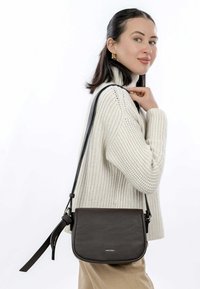 Borsa a tracolla in pelle marrone con superficie testurizzata, tracolla regolabile e branding sottile sul fronte, indossata con un maglione lavorato a maglia.