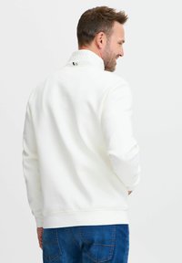 Felpa bianca con colletto alto, polsini e orlo a coste. Presenta un dettaglio del logo sul colletto. Abbinata a jeans blu.