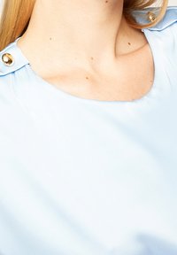 Blouse bleu clair avec un col rond et des boutons aux épaules. Le tissu semble lisse avec un léger brillant.