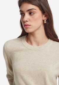 Beige tröja med rund halsringning, i en mjuk, strukturerad stickning. Modellen bär ett örhänge med guldaccent och har håret nedsläppt.