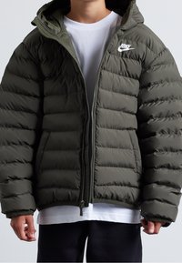 Grüne Daunenjacke mit gesteppter Oberfläche, Reißverschluss und Kapuze. Verfügt über ein kleines weißes Nike-Logo auf der linken Brust. Weicher Stoff.