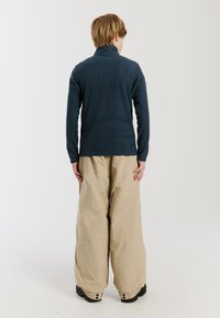 Pullover in pile blu navy con collo alto e maniche lunghe abbinato a pantaloni beige larghi. Il pullover presenta un'etichetta con logo sull'orlo.