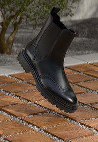 Must musta Chelsea saamiseks, millel on brogue detailid külgedel, tugeva tallaga ja peene disaini, rippumas tekstuurilisel kivikatte pinnal.