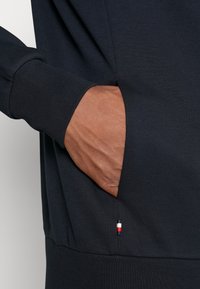 Main droite insérée dans la poche avant d'un sweat-shirt bleu marine foncé avec une petite étiquette rouge, blanche et bleue près de la couture de la poche.