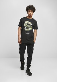 Svart t-skjorte i bomull med et grafisk design av gullgrillz og penger, kombinert med svarte cargo-bukser og svarte sneakers.