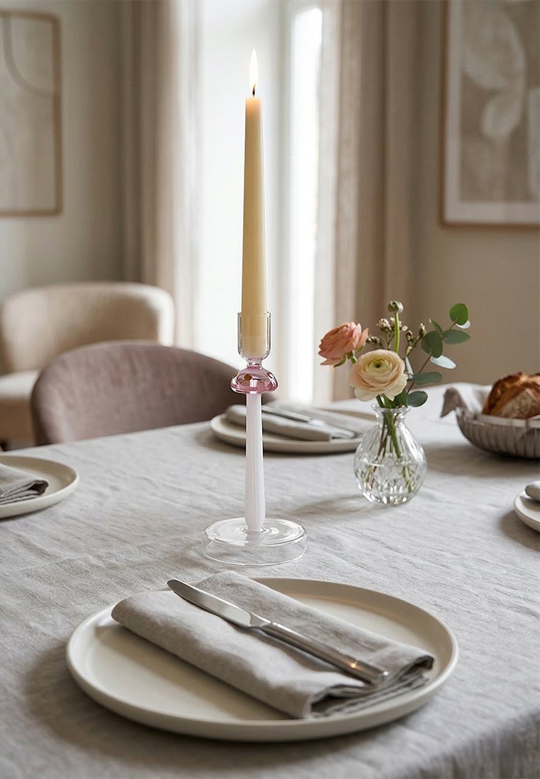 Zapalcana wysoka świeca w różowym szklanym świeczniku na stole jadalnym przykrytym lnianym obrusem, nakrytym talerzami, sztućcami, kwiatami i chlebem w pomieszczeniu o neutralnych tonach.