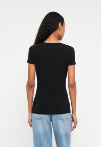 Kobieta z długimi kręconymi włosami, ubrana w dopasowaną czarną koszulkę z krótkim rękawem i jasnoniebieskie dżinsy, zwrócona plecami na białym tle.