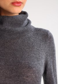 Maglione dolcevita grigio realizzato in morbido materiale a maglia, caratterizzato da un collo aderente e una texture liscia con sottili variazioni di luce.