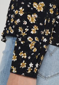 Tissu bleu marine foncé avec un motif floral jaune et blanc. Manches retroussées avec détail de boutons. Associé à un denim bleu clair.