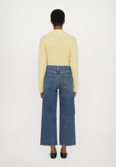 Geltona ilgomis rankovėmis palaidinė su apykakle, derinama su laisvai krentančiomis mėlynomis džinsinėmis kelnėmis. Juodus lygus batus užbaigia aprangą.