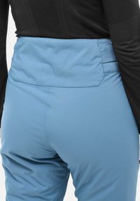 Pantalon bleu clair avec une texture lisse et une taille élastique, doté de poches arrière et de détails de couture. Porté avec un haut noir.