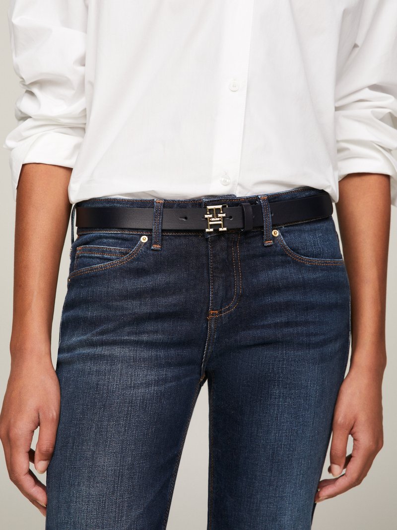 Pessoa a usar jeans azul-escuro com costura laranja, uma camisa branca de manga longa com botões, e um cinto de couro preto com uma fivela dourada em forma de "H".