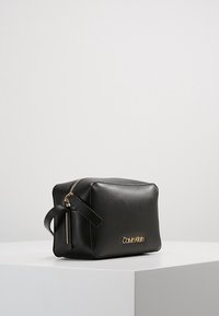 Černá kožená taška obdélníkového tvaru se zlatým zipem a logem, s postranním úchytem pro snadné nošení. Hladký povrch, minimalistický design.