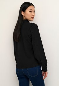 Blouse noire texturée avec manches longues bouffantes, col montant et poignets à boutons, associée à un jean bleu ajusté.
