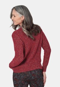 Roter strukturierter Pullover mit lockerer Passform und geripptem Saum, kombiniert mit dunkel gemusterten Hosen, von hinten in einem Winkel präsentiert.