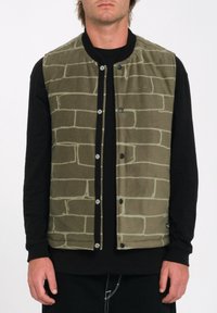 Chaleco de forro polar verde con diseño de ladrillos, cierres de presión y cuello negro; llevado sobre una camiseta de manga larga negra.