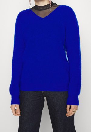 Pull bleu duveteux porté sur un top noir transparent, associé à un jean foncé, montrant des mains avec des bagues et de longues boucles d’oreilles pendantes.