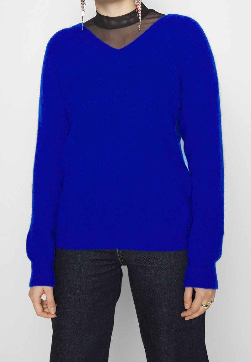 Pull bleu duveteux porté sur un top noir transparent, associé à un jean foncé, montrant des mains avec des bagues et de longues boucles d’oreilles pendantes.